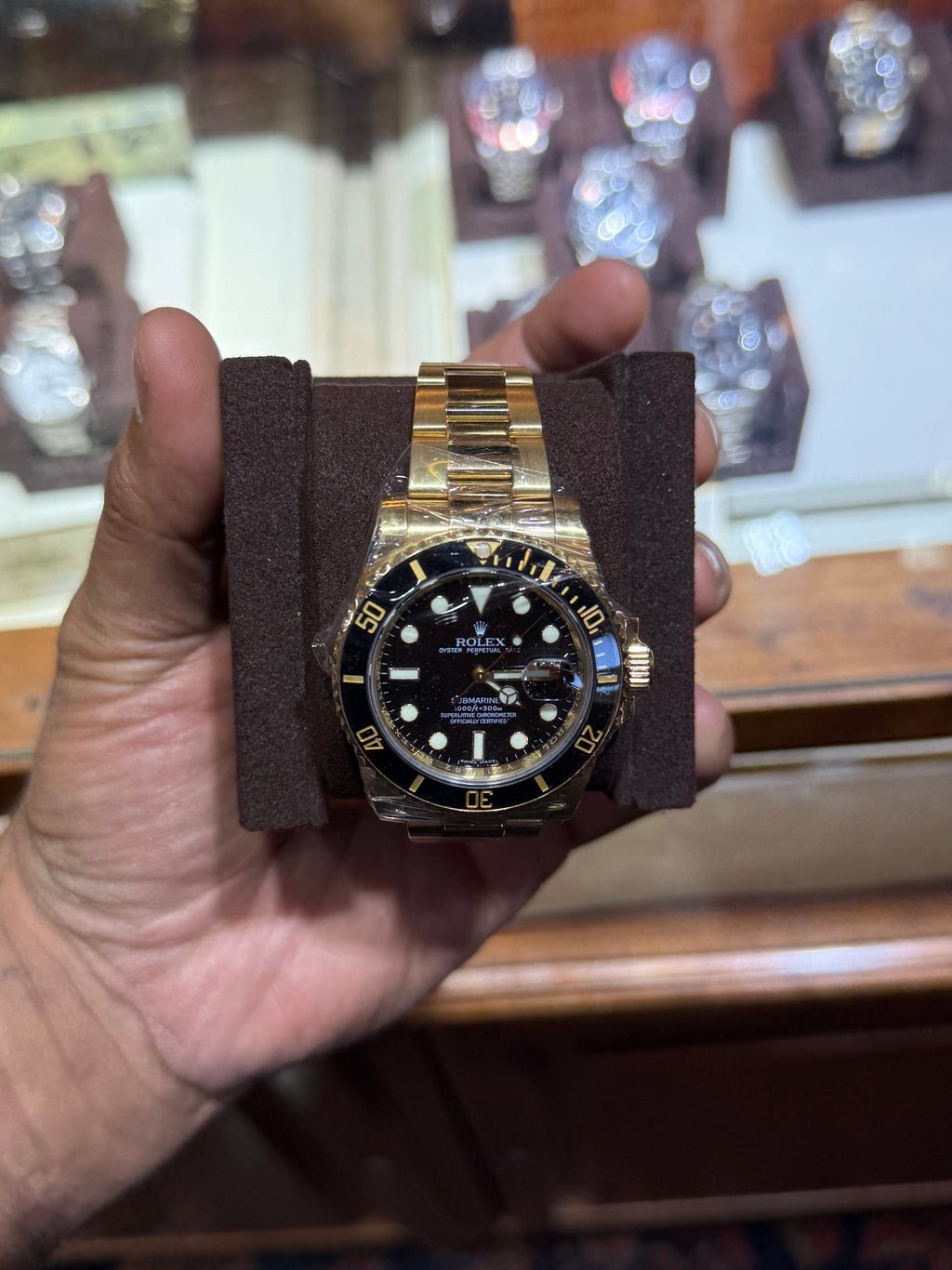 A gold-tone Rolex Submariner watch with a black dial and black bezel, held in a person's hand on a dark brown display cushion. The background shows a wooden watch display case with other watches inside.