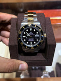 A Rolex Submariner Date watch with a two-tone gold and silver stainless steel bracelet, black dial, and black bezel with gold markings, displayed in a dark brown velvet watch box held by a hand.