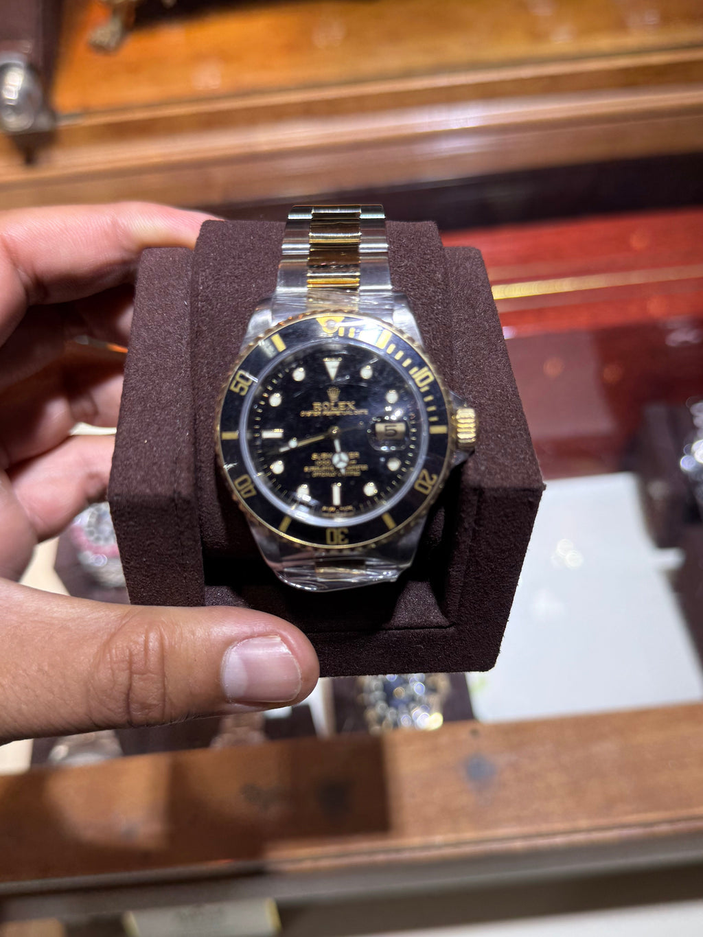 A Rolex Submariner Date watch with a black dial and gold-tone bezel, presented in a dark brown velvet watch box held by a hand.