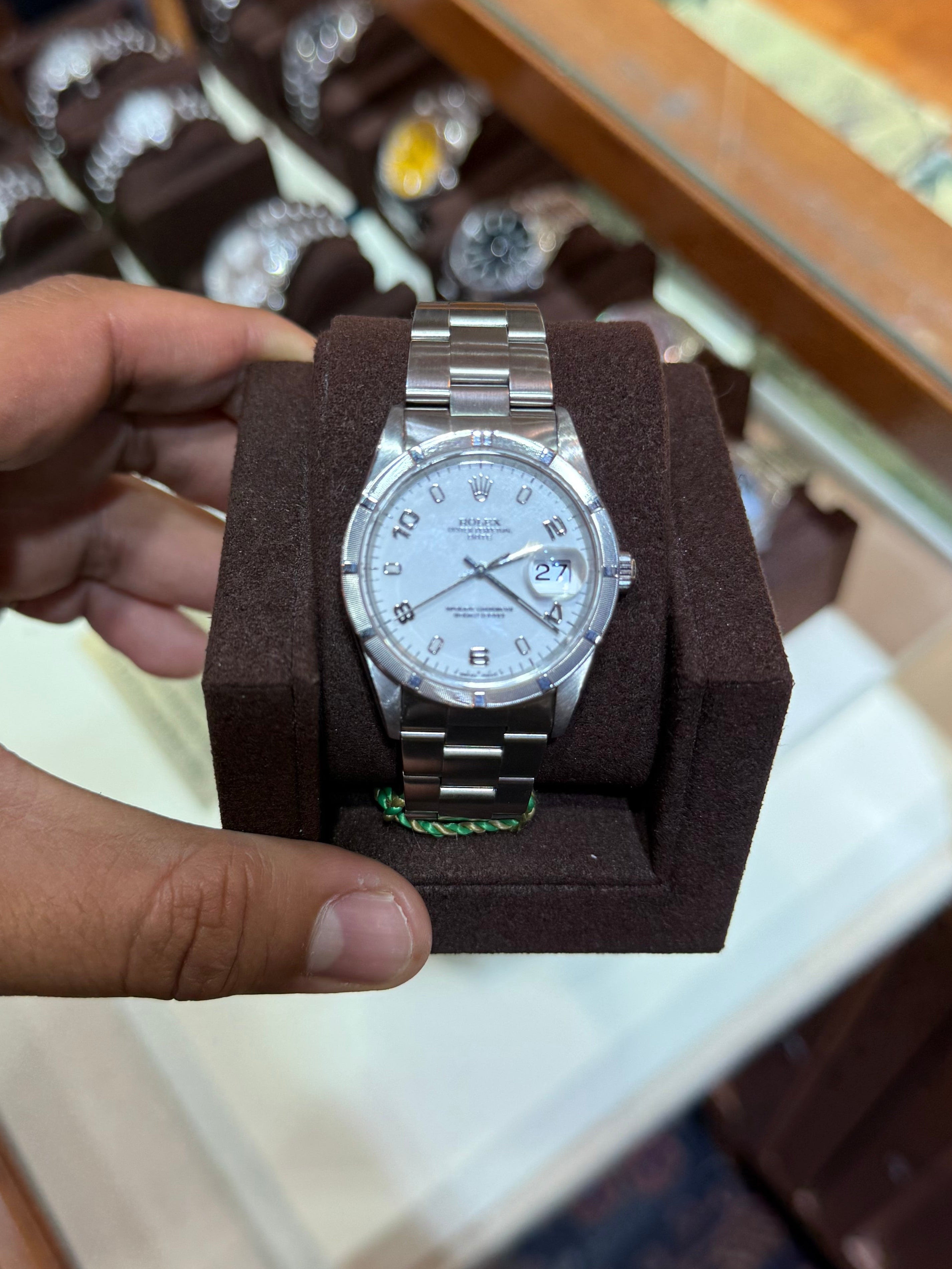 A silver stainless steel wristwatch with a white dial and date window, displayed inside a dark brown rectangular presentation box held by a hand.