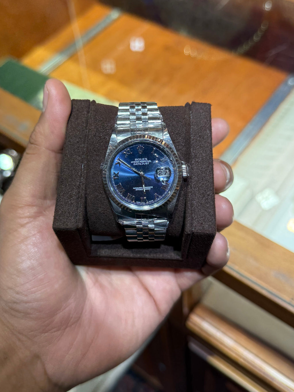 A silver-tone stainless steel wristwatch with a blue dial and matching blue strap, displayed on a dark brown rectangular stand held in a person's hand. The background shows a warm, blurred wooden display case or cabinet.