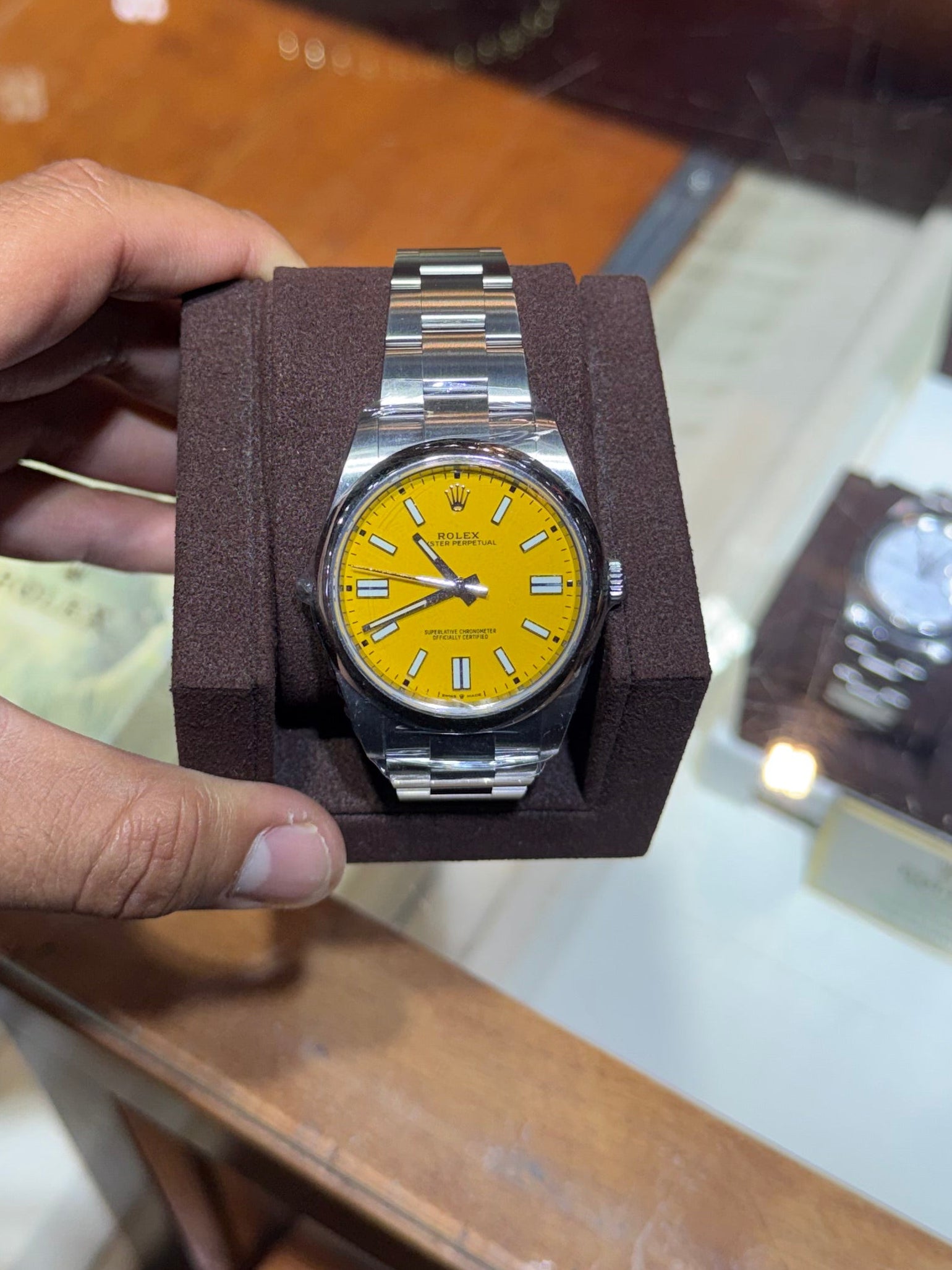 A wristwatch with a bright yellow dial and silver metal bracelet displayed on a dark brown cushion, held by a hand in front of a wooden watch display case.