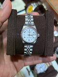 A silver-tone stainless steel watch with a white dial and matching metal bracelet, displayed on a dark brown cushion inside a person's hand, with a watch display case in the blurred background.