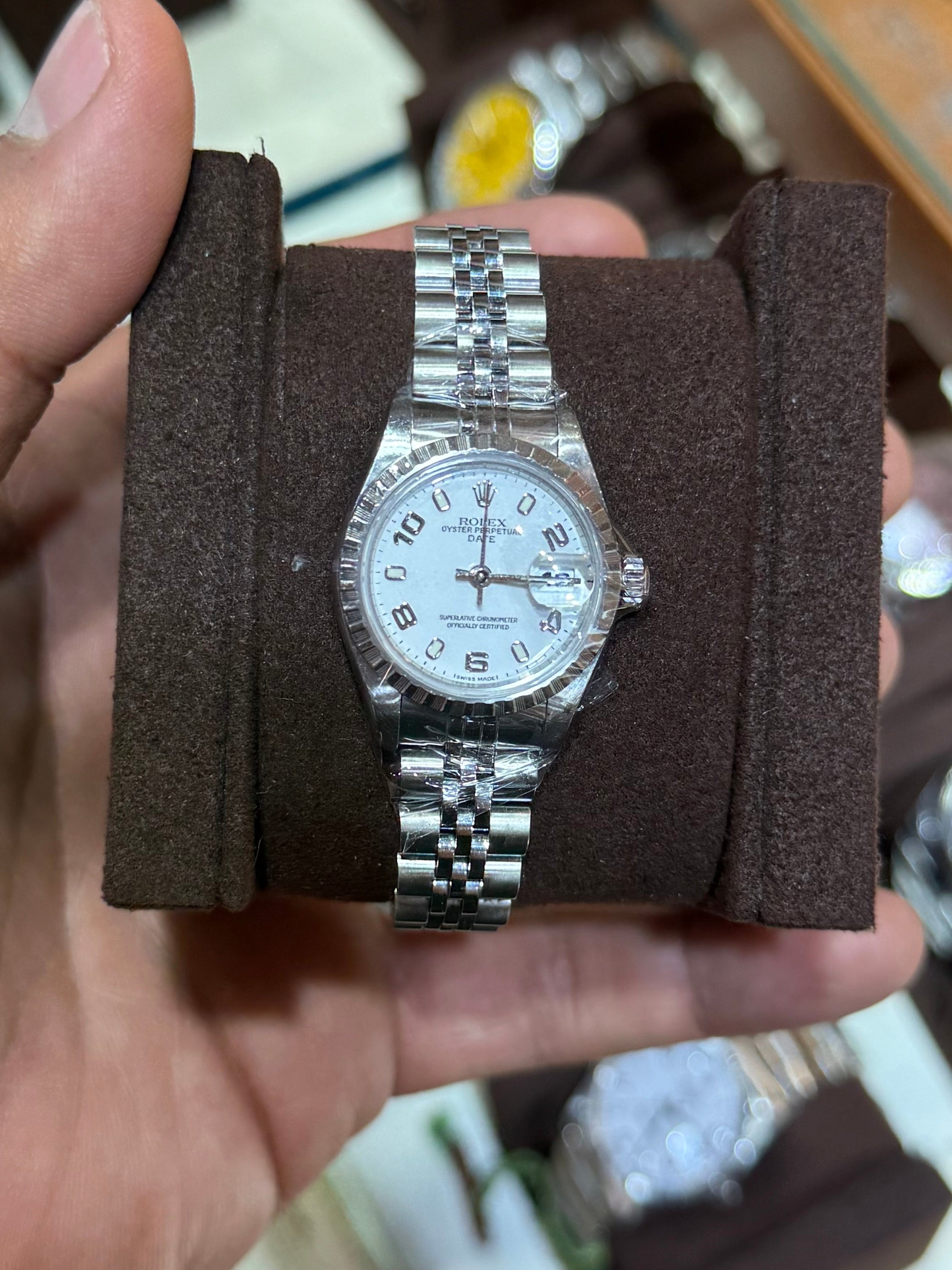 A silver-tone stainless steel watch with a white dial and matching metal bracelet, displayed on a dark brown cushion inside a person's hand, with a watch display case in the blurred background.