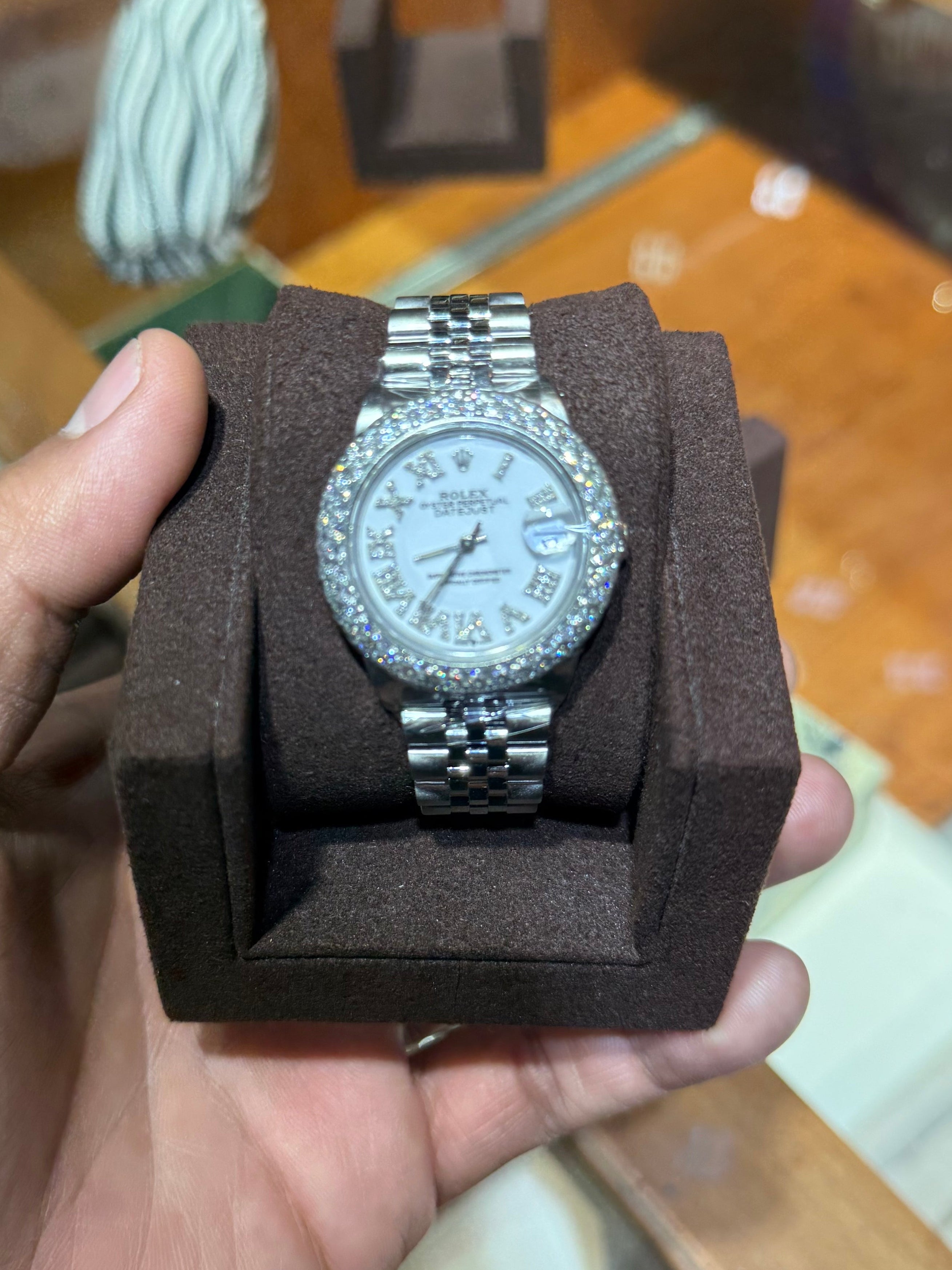A silver-tone stainless steel watch with a diamond-encrusted bezel and silver-tone link bracelet, displayed on a dark brown velvet watch cushion held by a hand. The background shows a wooden display case in a shop.