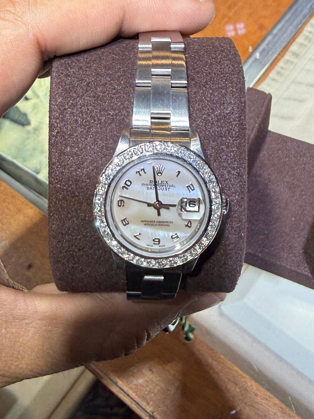 A silver-tone stainless steel wristwatch with a white dial and a bezel set with clear stones, held in a person's hand on a brown display cushion inside a watch store.