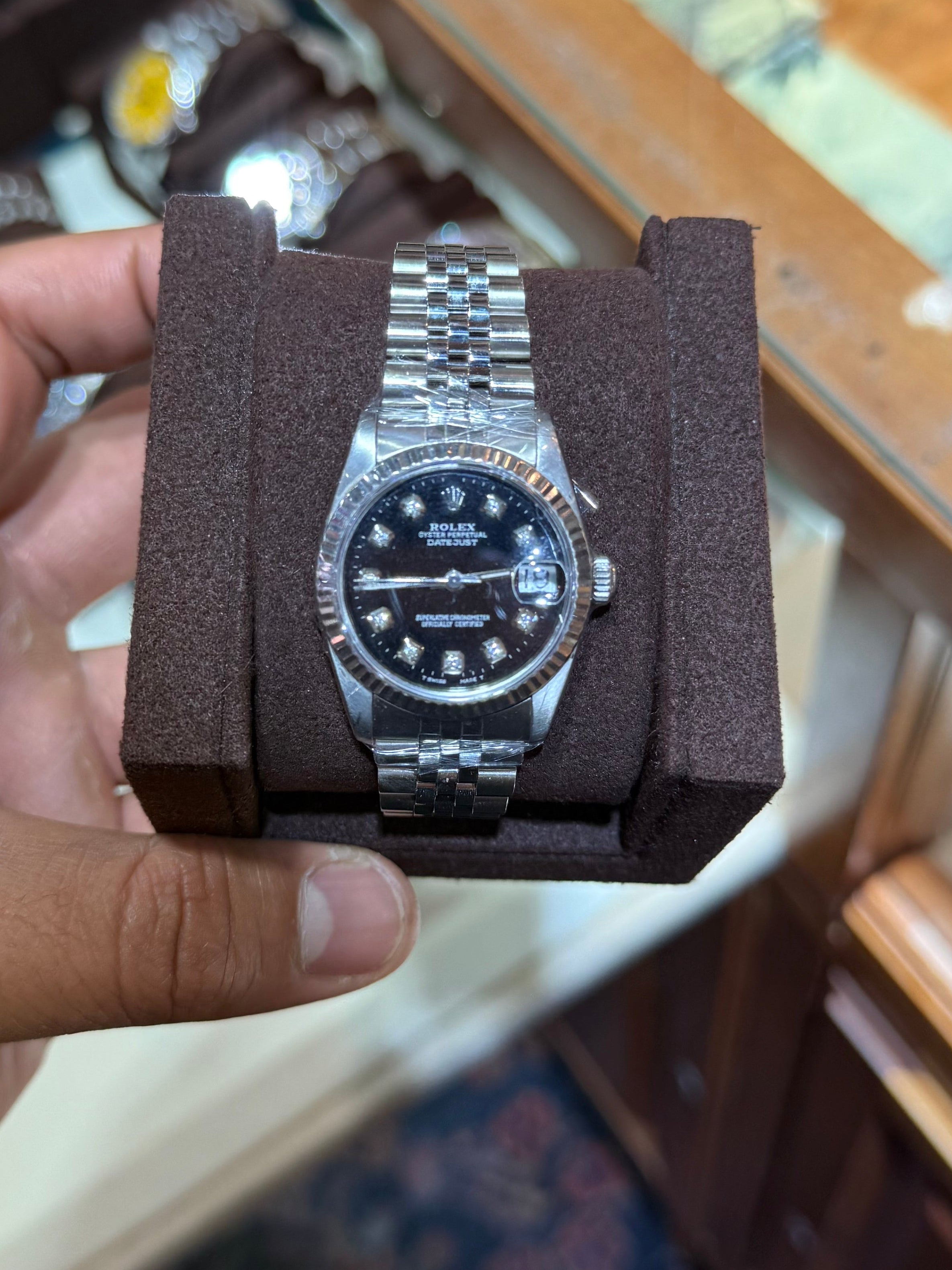 A silver stainless steel wristwatch with a black dial and matching steel bracelet, displayed inside a dark brown rectangular presentation box held by a person's hand.