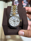 A gold and silver stainless steel watch with a white dial and crystal hour markers, displayed on a brown velvet watch cushion and held by a hand in front of a blurred watch display case.