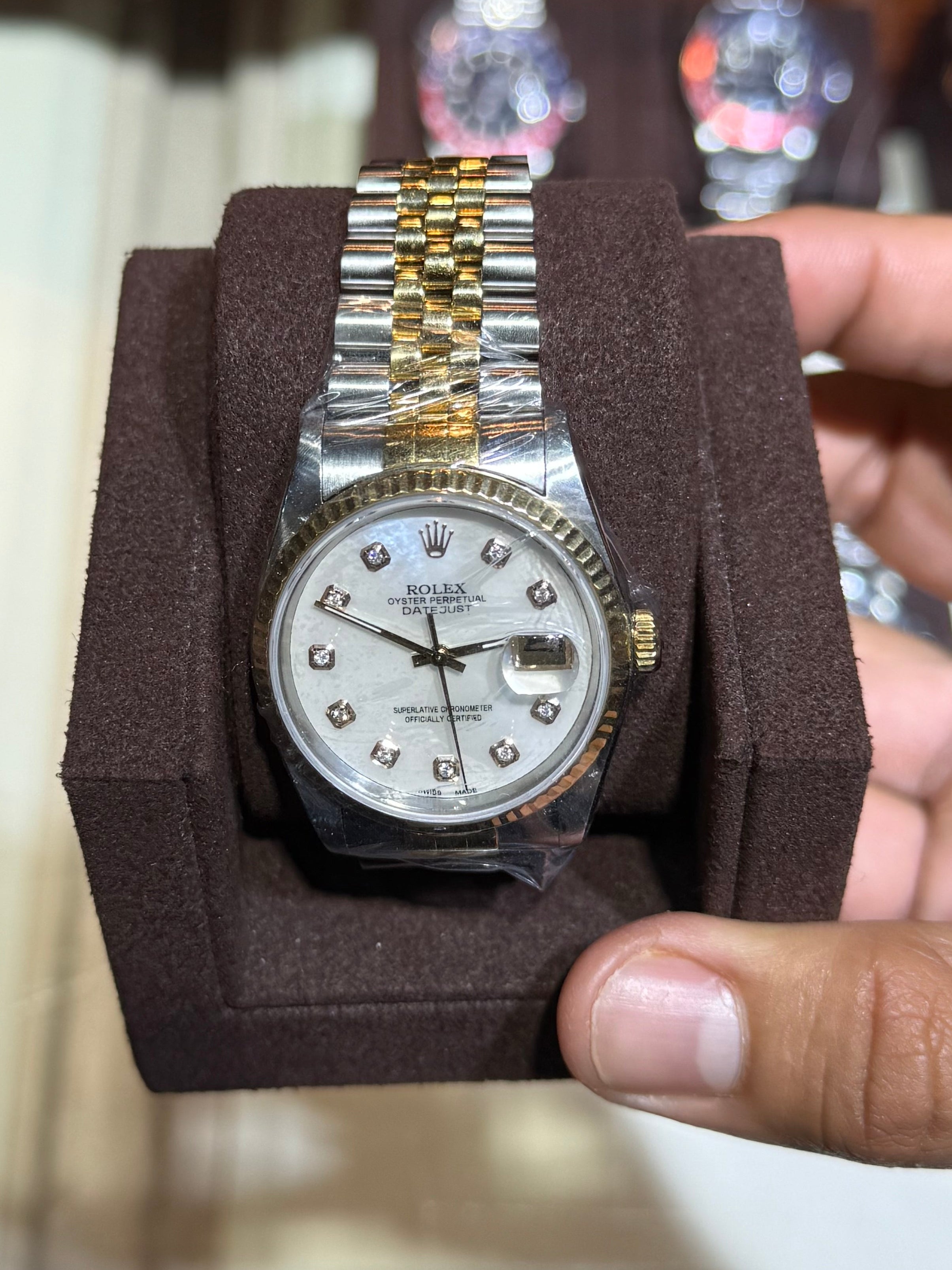 A gold and silver stainless steel watch with a white dial and crystal hour markers, displayed on a brown velvet watch cushion and held by a hand in front of a blurred watch display case.
