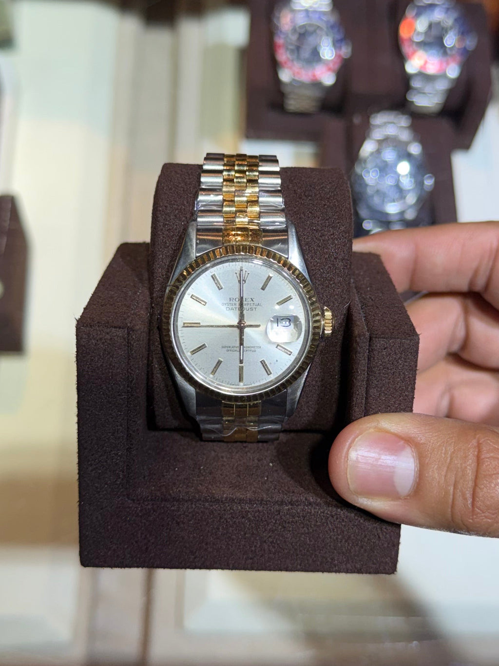A gold and stainless steel wristwatch with a white dial is held in a dark brown display stand by a person's hand, with a glass display case of other watches in the background.