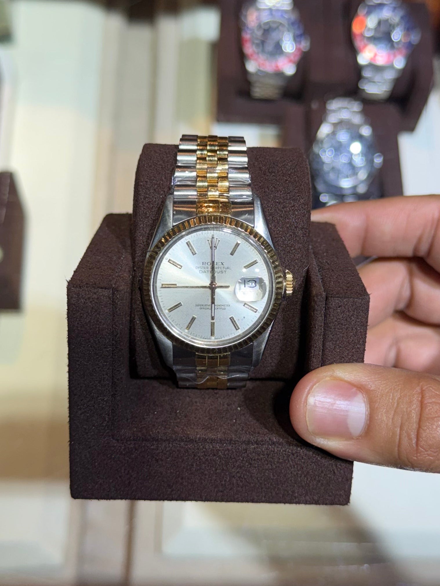 A gold and stainless steel wristwatch with a white dial is held in a dark brown display stand by a person's hand, with a glass display case of other watches in the background.
