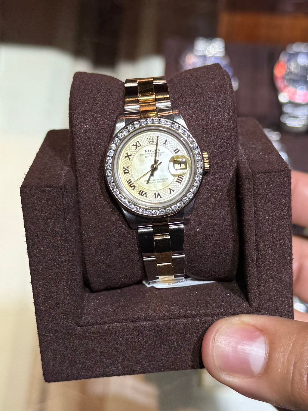 A gold and stainless steel wristwatch with a cream dial is displayed on a brown velvet watch cushion, held by a hand in front of a vintage watch display case.