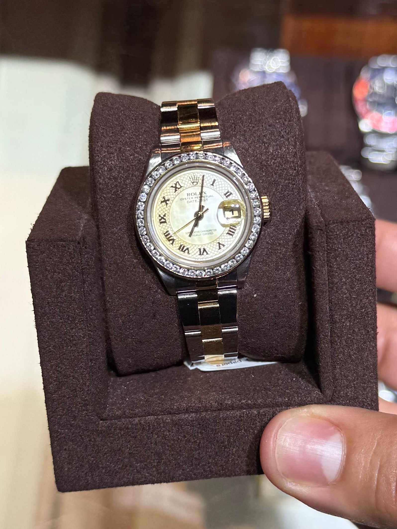A gold and stainless steel wristwatch with a cream dial is displayed on a brown velvet watch cushion, held by a hand in front of a vintage watch display case.
