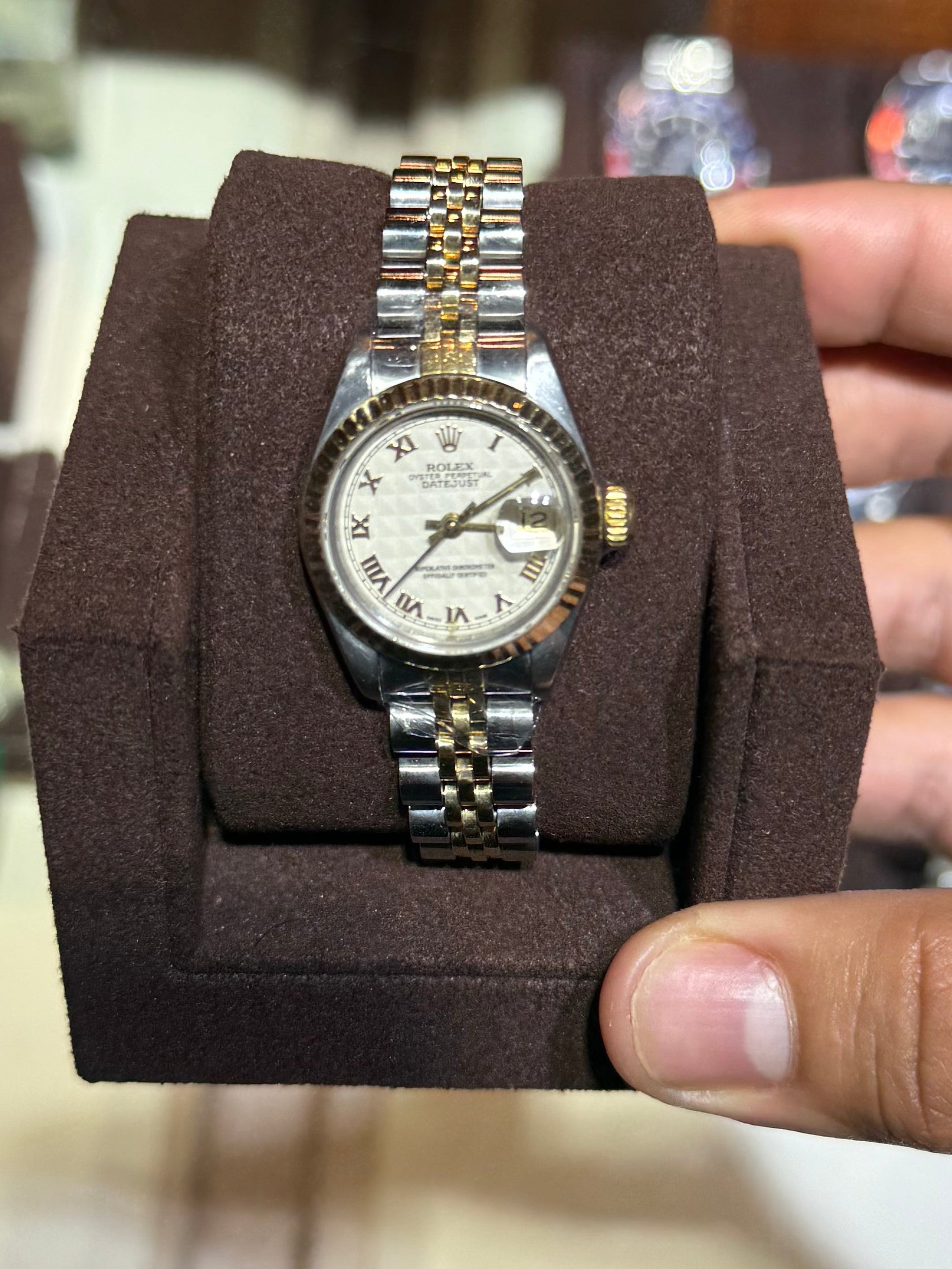 A vintage two-tone wristwatch with a white dial and silver-and-gold metal bracelet, displayed on a dark brown cushion and held by a hand in front of a watch display case.