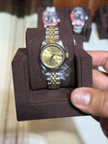 A gold-tone and stainless steel two-tone wristwatch with a gold dial, displayed on a dark brown velvet watch cushion and held by a hand in front of a glass display case.
