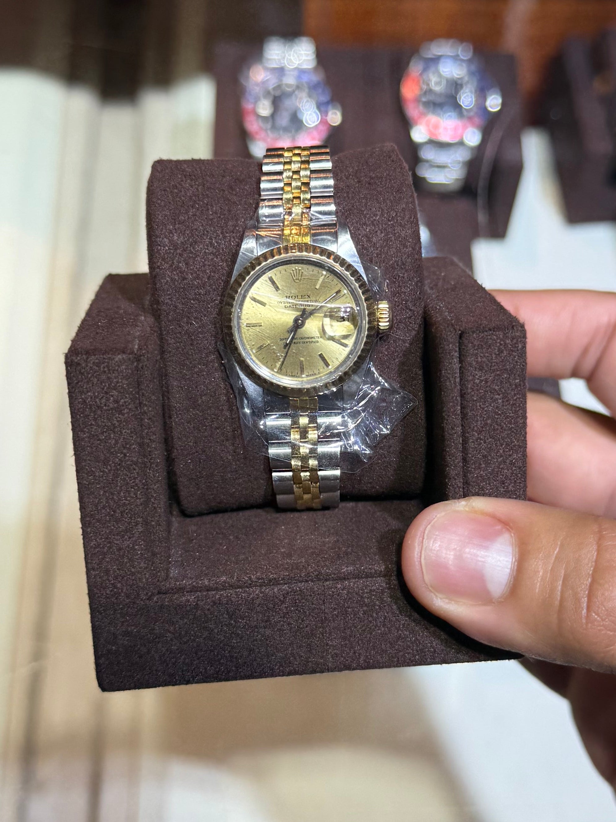A gold-tone and stainless steel two-tone wristwatch with a gold dial, displayed on a dark brown velvet watch cushion and held by a hand in front of a glass display case.