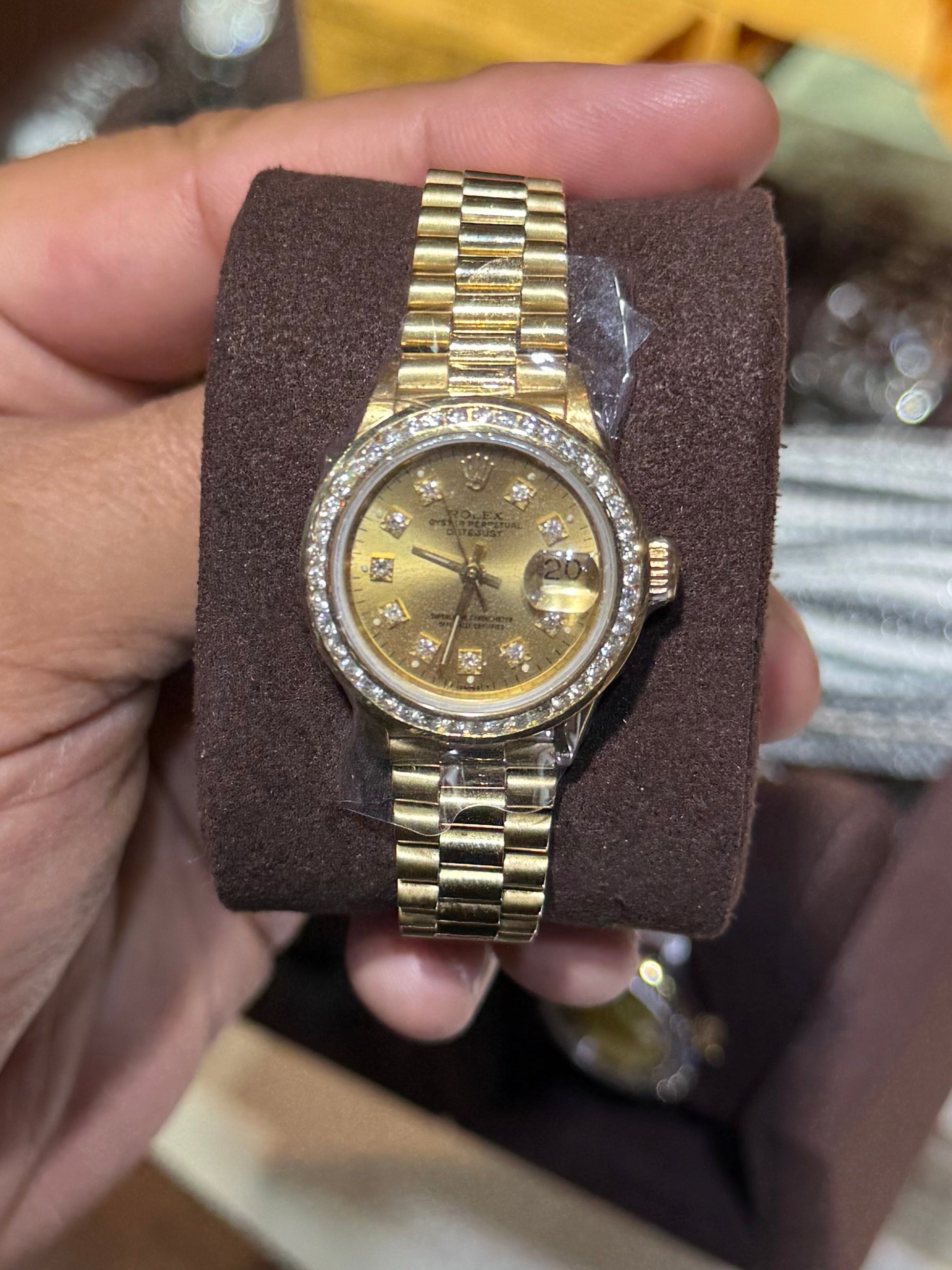 A gold-tone wristwatch with a matching metal link bracelet and a bezel set with small clear stones, held in a person's hand over a brown display case.