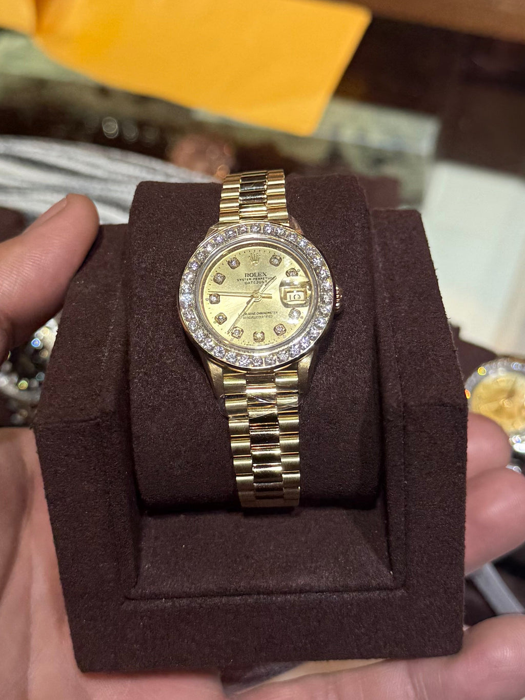 A gold-tone stainless steel watch with a gold dial and a diamond-encrusted bezel is displayed on a brown velvet watch stand, held in a person's hand inside a watch shop.