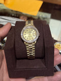A gold-tone stainless steel watch with a gold dial and a diamond-encrusted bezel is displayed on a brown velvet watch stand, held in a person's hand inside a watch shop.