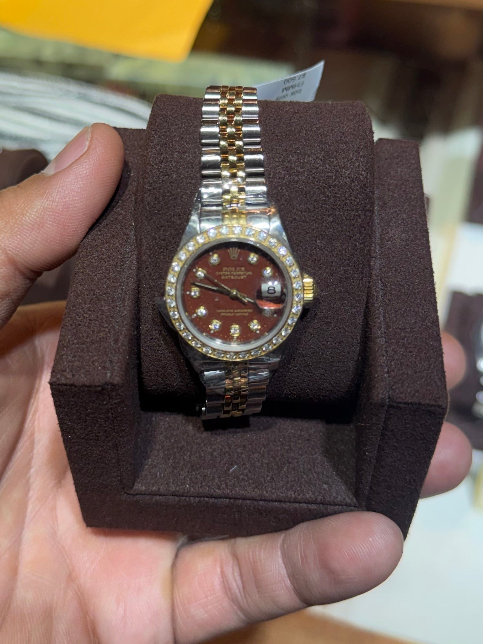 A gold and silver-tone wristwatch with a brown dial is displayed on a dark brown cushion held by a person's hand in a watch store.