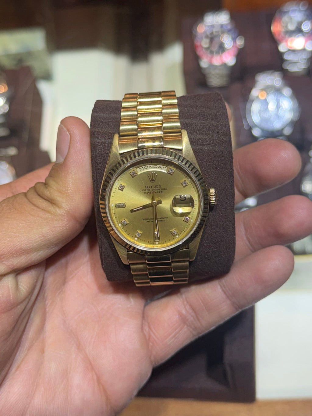 A gold-tone wristwatch with a matching metal bracelet is held in a person's hand in front of a wooden watch display case.