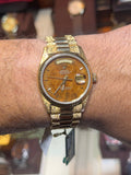 A gold-tone wristwatch with a brown dial and matching metal bracelet worn on a person's wrist, photographed in a watch display setting with blurred clocks and other watches in the background.