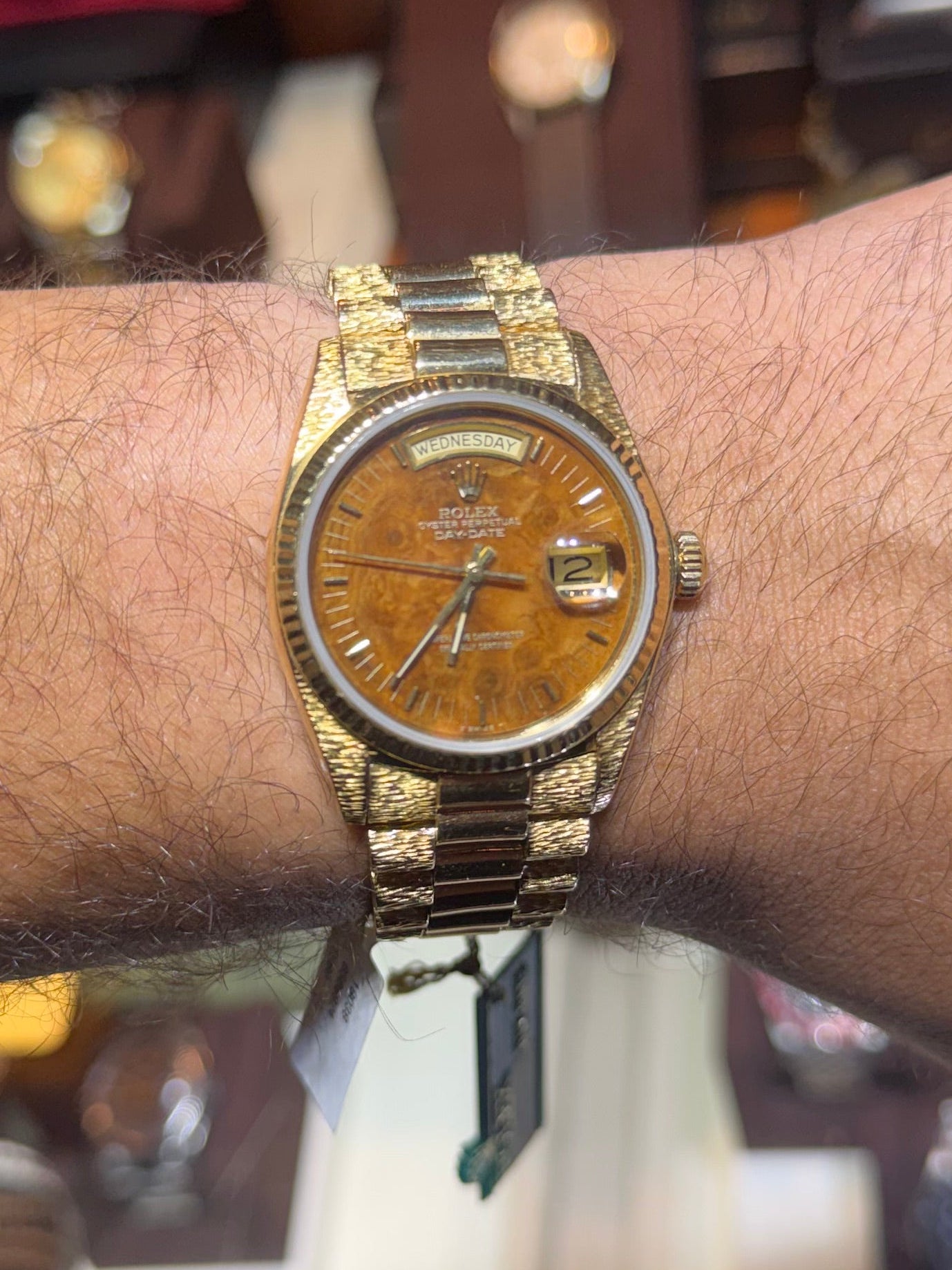A gold-tone wristwatch with a brown dial and matching metal bracelet worn on a person's wrist, photographed in a watch display setting with blurred clocks and other watches in the background.
