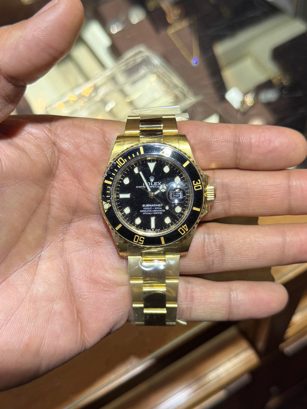 A gold-tone stainless steel dive watch with a black dial and black bezel, held in a person's hand in front of a wooden watch display case.