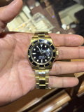 A gold-tone stainless steel dive watch with a black dial and black bezel, held in a person's hand in front of a wooden watch display case.