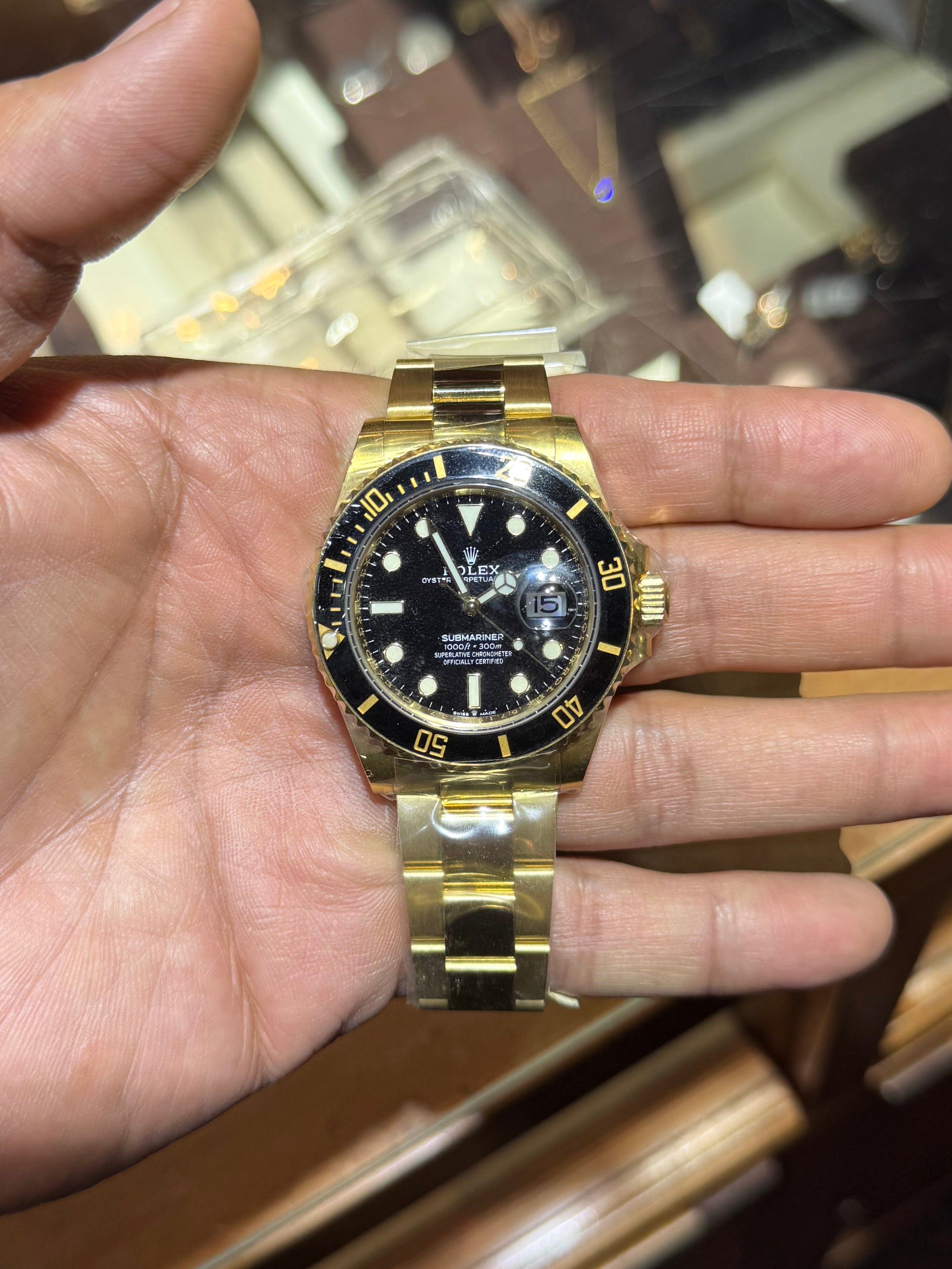 A gold-tone stainless steel dive watch with a black dial and black bezel, held in a person's hand in front of a wooden watch display case.
