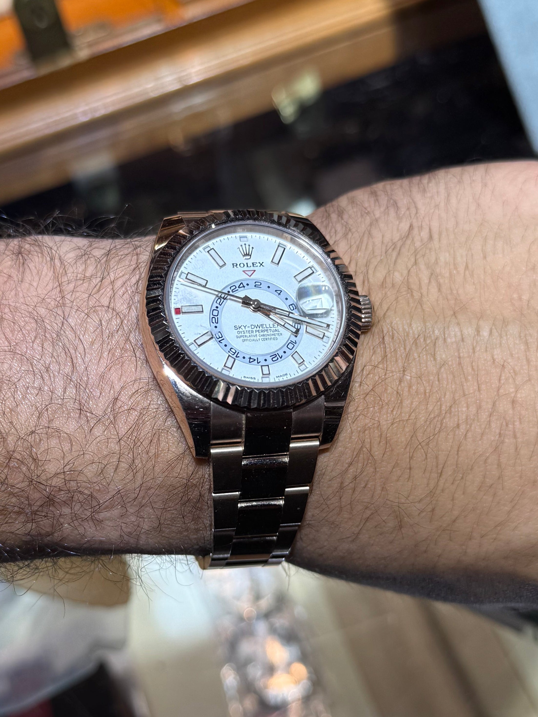 A wristwatch with a rose gold-toned metal bracelet and case, featuring a white dial with blue accents, shown worn on a person's wrist in a watch store.