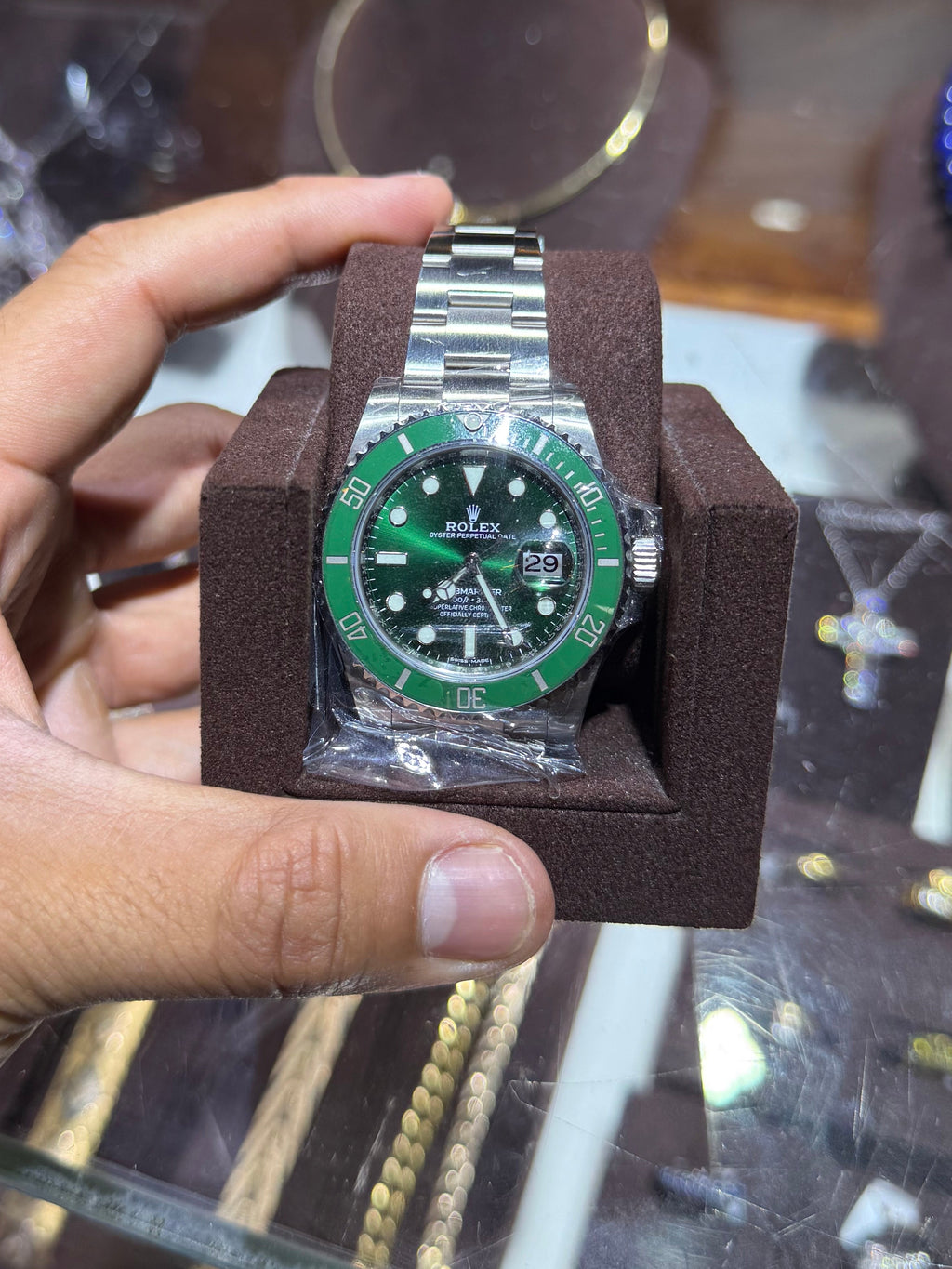 A silver-tone stainless steel watch with a green dial and green bezel, held in a brown display cushion by a person's hand in front of a jewelry display case.