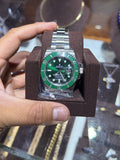 A silver-tone stainless steel watch with a green dial and green bezel, held in a brown display cushion by a person's hand in front of a jewelry display case.